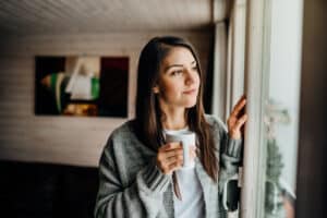 Woman holding mug looks out the window, trying to avoid depression symptoms.